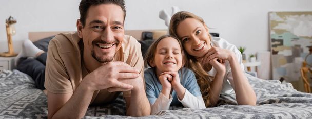 portrait of happy parents and kid looking at camera while resting in bedroom, banner  - 写真・画像
