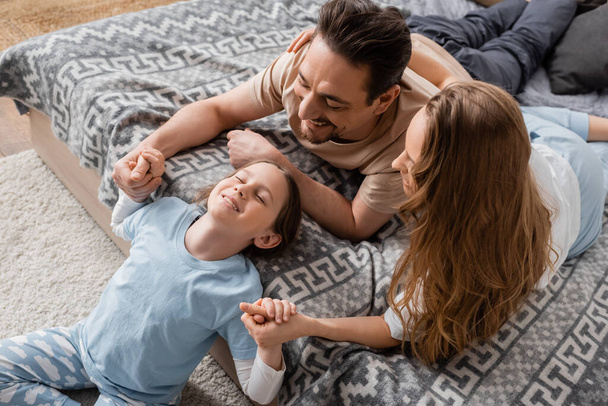 высокий угол зрения положительный ребенок сидит на ковре рядом счастливые родители на кровати  - Фото, изображение