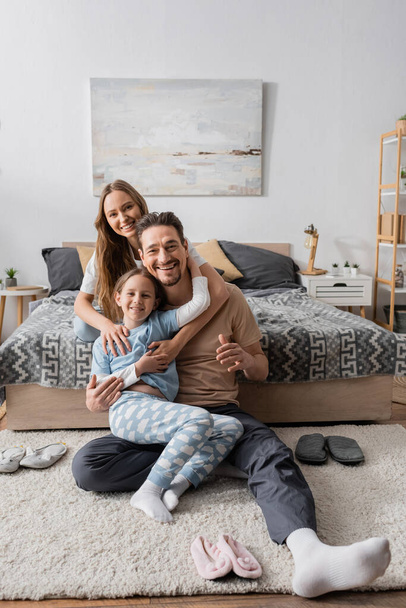 cheerful kid sitting with happy father on carpet around slippers near happy mother on bed  - Foto, Bild