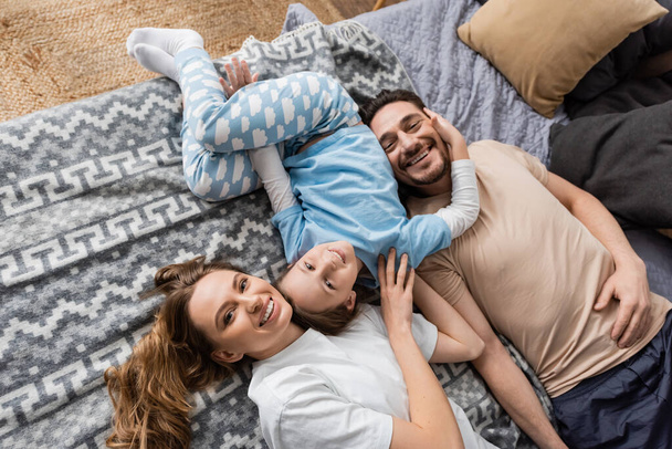 top view of positive parents and cheerful kid lying on bed at home  - Fotó, kép