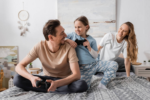KYIV, UKRAINE - NOVEMBER 28, 2022: cheerful father and child playing video game near wife in bedroom  - Foto, Bild