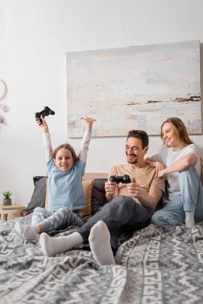 KYIV, UKRAINE - NOVEMBER 28, 2022: cheerful woman leaning on shoulder of husband playing video game with excited daughter  - 写真・画像