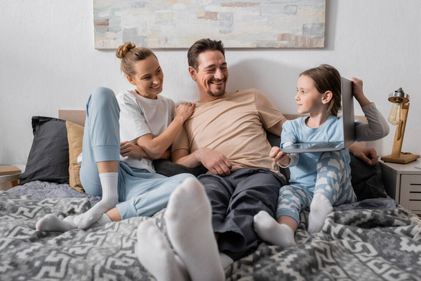 cheerful parents looking at happy kid holding laptop in bedroom  - Foto, Imagem