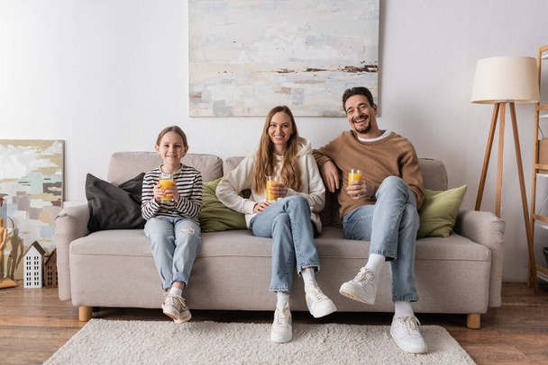full length των χαρούμενων οικογένεια εκμετάλλευση ποτήρια χυμό πορτοκαλιού και κάθεται στον καναπέ  - Φωτογραφία, εικόνα