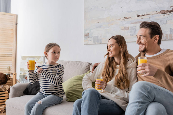 веселые родители, держащие стаканы апельсинового сока и смотрящие на дочь, сидя на диване  - Фото, изображение