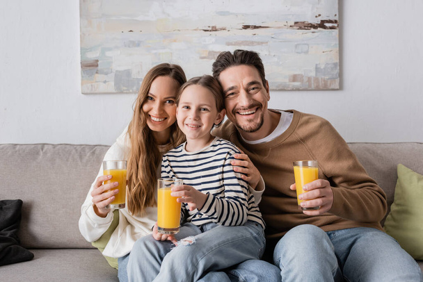 счастливая дочь сидит рядом с позитивными родителями, держа стаканы апельсинового сока  - Фото, изображение