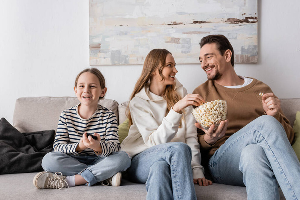 cheerful parents eating popcorn near happy daughter with remote controller  - Foto, immagini