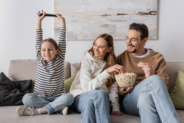 cheerful parents eating popcorn near excited daughter with remote controller  - Foto, imagen
