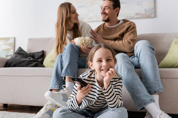 cheerful kid with remote controller watching movie near parents sitting with popcorn on blurred background  - Foto, Imagem