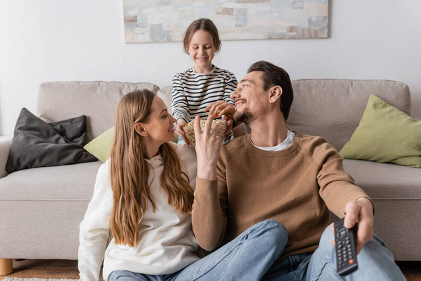 happy man holding remote controller and reaching popcorn near wife and cheerful daughter on sofa  - Foto, Bild