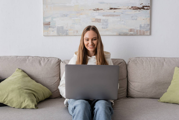 χαρούμενη γυναίκα σε casual ρούχα χρησιμοποιώντας φορητό υπολογιστή, ενώ κάθεται στον καναπέ  - Φωτογραφία, εικόνα