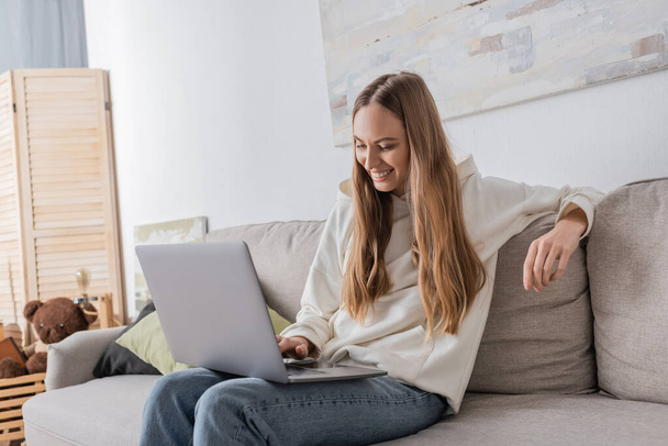 positive freelancer in casual clothes using laptop while sitting on couch  - Photo, image