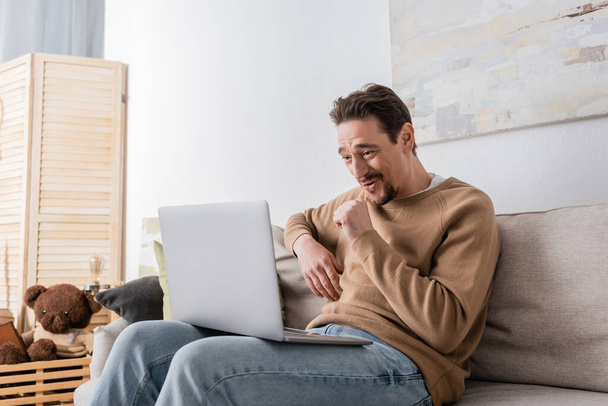 бородатый человек смотрит фильм на ноутбуке, сидя на диване в гостиной  - Фото, изображение