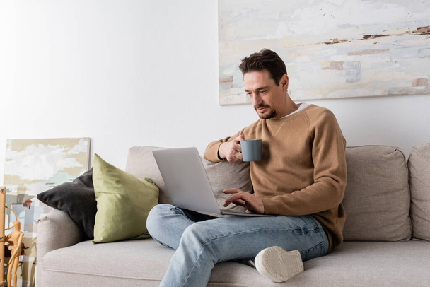 бородатий чоловік використовує ноутбук, тримаючи чашку і сидячи на дивані у вітальні
  - Фото, зображення