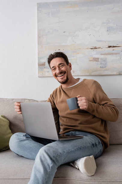 радостный человек с помощью ноутбука, держа чашку и сидя на диване в гостиной  - Фото, изображение