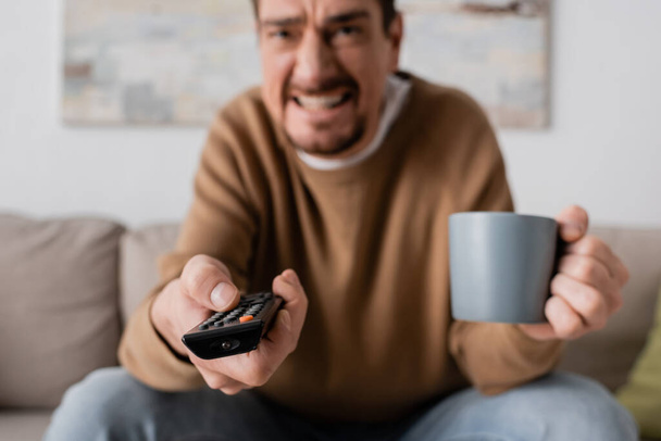 angry man holding remote controller and cup of coffee in living room  - Fotoğraf, Görsel