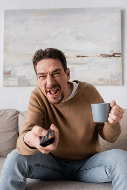 напряженный человек держа пульт дистанционного управления и чашку кофе в гостиной  - Фото, изображение