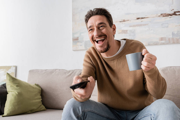 amazed man holding remote controller and cup of coffee in living room  - Fotó, kép