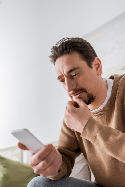 low angle view of bearded man in beige jumper messaging on smartphone at home  - Fotó, kép