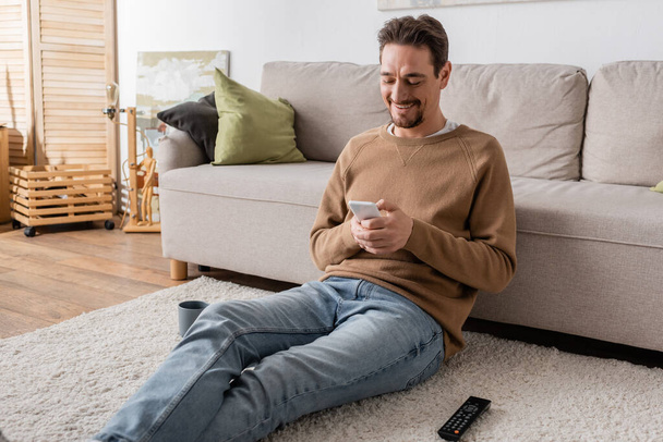 家でカーペットの上に座っている間にスマートフォンでベージュのジャンパーメッセージで幸せな男  - 写真・画像