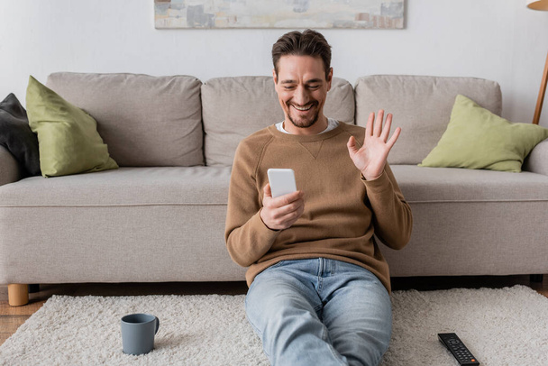 家でビデオ通話をしている間にスマートフォンで手を振っているベージュジャンパーの陽気な男  - 写真・画像