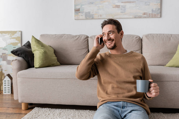 cheerful man in beige jumper talking on smartphone while sitting with cup of coffee at home  - Fotoğraf, Görsel