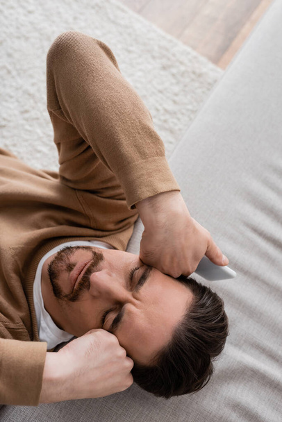 вид сверху на уставшего человека в бежевом свитере, держащего мобильный телефон и опирающегося на диван  - Фото, изображение