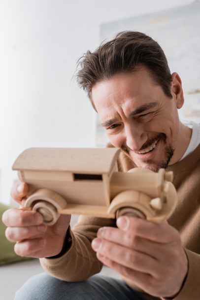 веселий чоловік посміхається, дивлячись на дерев'яну іграшку автомобіля у вітальні
  - Фото, зображення