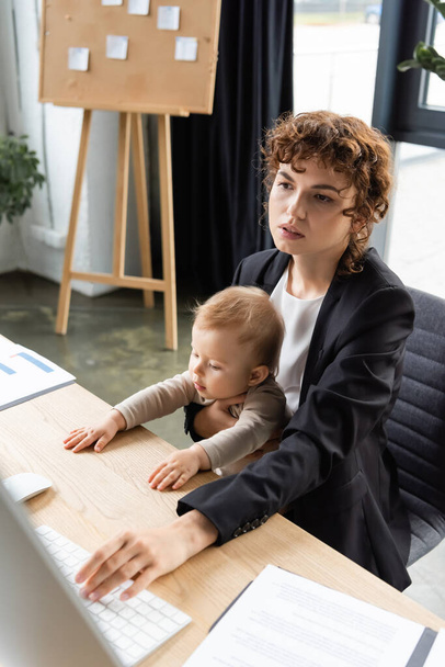 businesswoman sitting with little daughter at workplace and using computer near documents on desk - Foto, Imagen
