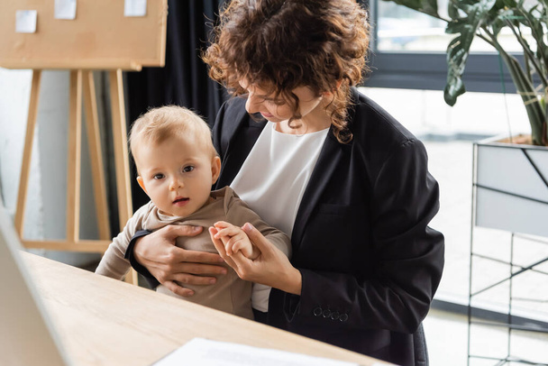 businesswoman in black blazer holding hand of toddler child while sitting at workplace in office - Foto, afbeelding