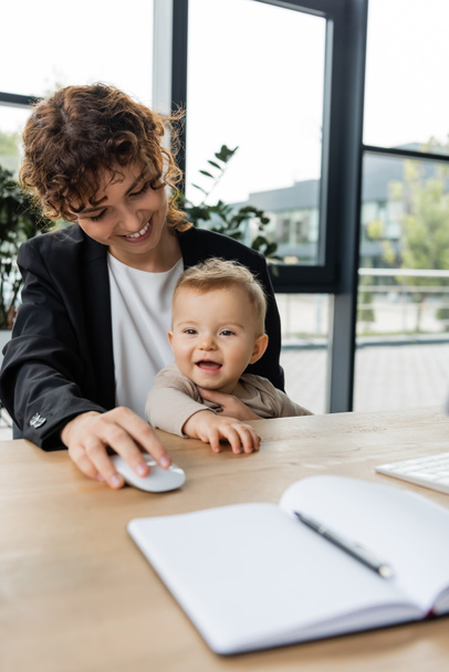 smiling businesswoman holding computer mouse near little cheerful daughter and blank notebook on blurred foreground - Foto, Imagem