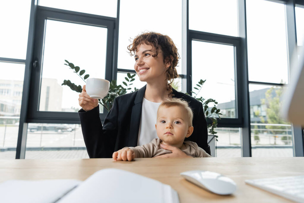 щаслива бізнес-леді, сидячи з маленькою дочкою і чашкою кави біля розмитої комп'ютерної миші і ноутбука на робочому столі
 - Фото, зображення