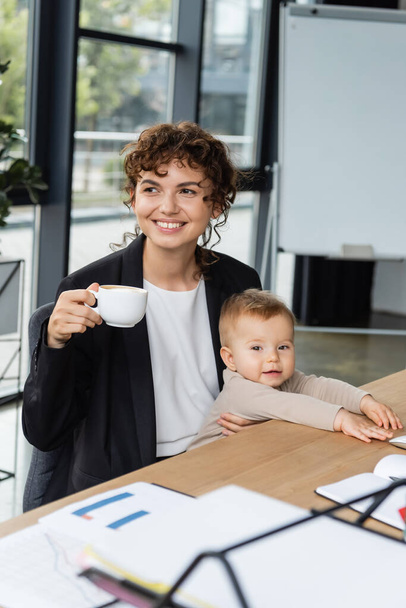 オフィスの職場で幼児の子供とコーヒーカップと一緒に座っていると笑顔の実業家 - 写真・画像
