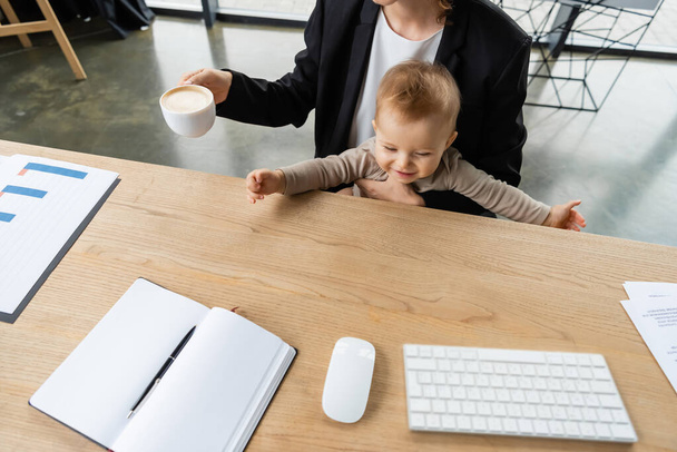 high angle view of cheerful baby near mother with coffee cup and computer mouse with keyboard and blank notebook on office desk - Photo, image