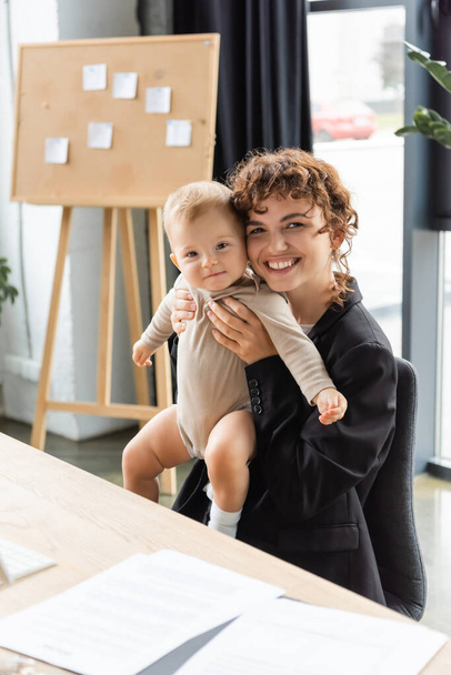 cheerful businesswoman holding little daughter in romper while sitting at work desk near blurred documents - Fotó, kép