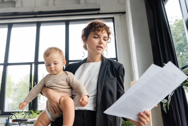 office manager in black blazer looking at papers while holding toddler baby in office - Photo, Image