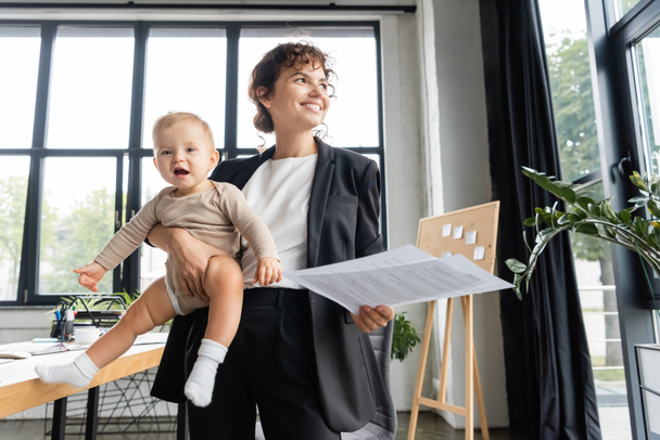 happy businesswoman in black suit looking away while standing with baby and documents in office - Zdjęcie, obraz