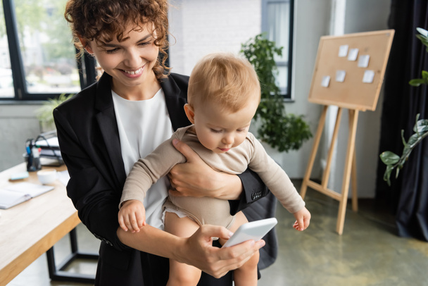 businesswoman in black blazer holding little daughter and taking selfie on mobile phone in office - Фото, зображення