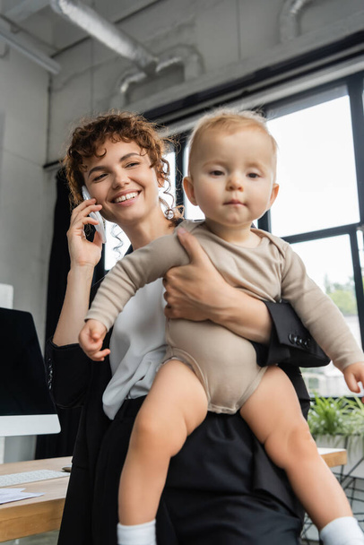 positive businesswoman talking on smartphone and holding toddler child in office - Foto, Imagen