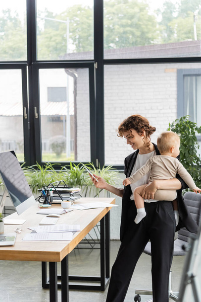 happy businesswoman standing with smartphone and baby near work desk with documents and computer monitor with blank screen - Fotografie, Obrázek