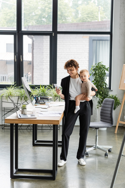 positive businesswoman holding smartphone and little child near work desk in modern office - Foto, afbeelding