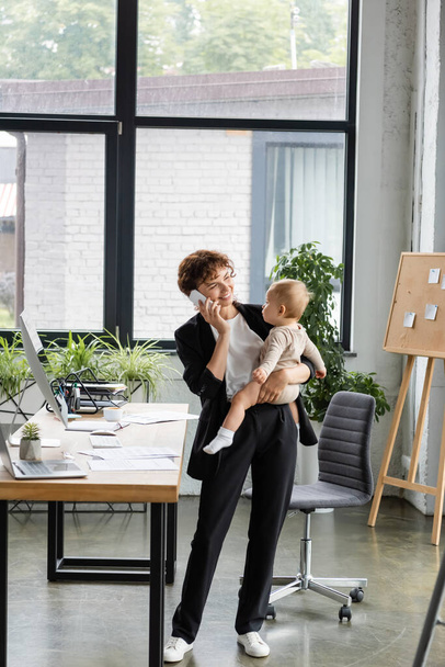 smiling businesswoman in black suit holding little daughter and talking on mobile phone in modern office - Foto, afbeelding