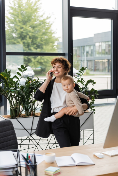 happy businesswoman looking away while holding baby and talking on mobile phone in modern office - Foto, afbeelding