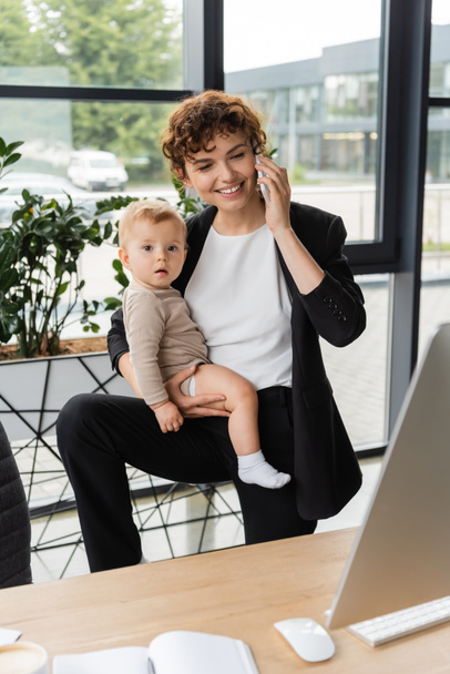 joyful businesswoman holding toddler daughter and talking on smartphone near computer monitor in office - Photo, image