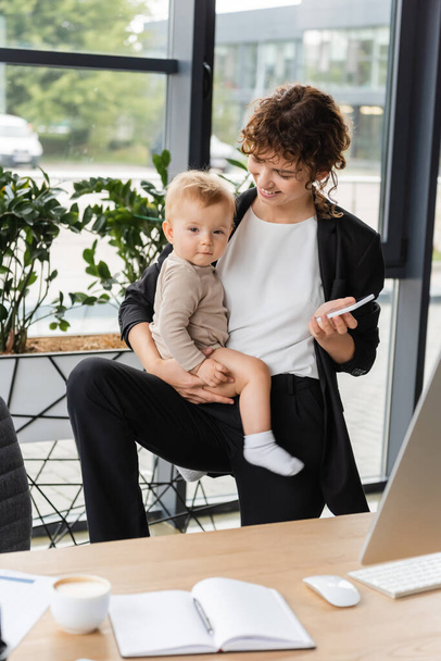 smiling businesswoman with baby and mobile phone near blurred notebook and coffee cup on work desk - Фото, зображення