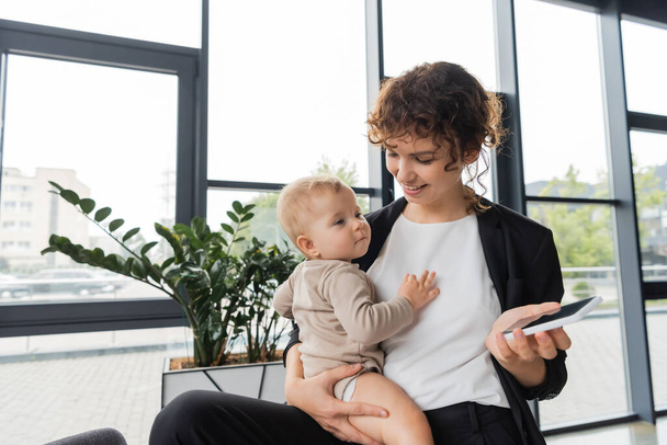 ευχαριστημένη επιχειρηματίας κρατώντας το μωρό και το κινητό τηλέφωνο με λευκή οθόνη κοντά σε πράσινα φυτά στο γραφείο - Φωτογραφία, εικόνα