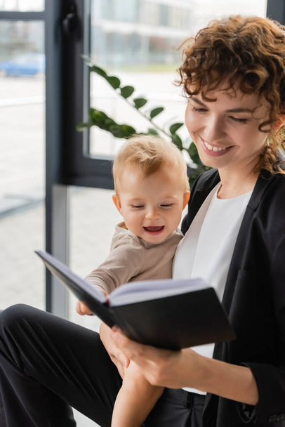 smiling businesswoman holding cheerful toddler daughter and looking in notebook in office - Foto, Imagen