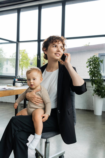 positive businesswoman looking away while sitting with baby in office and talking on smartphone - Zdjęcie, obraz