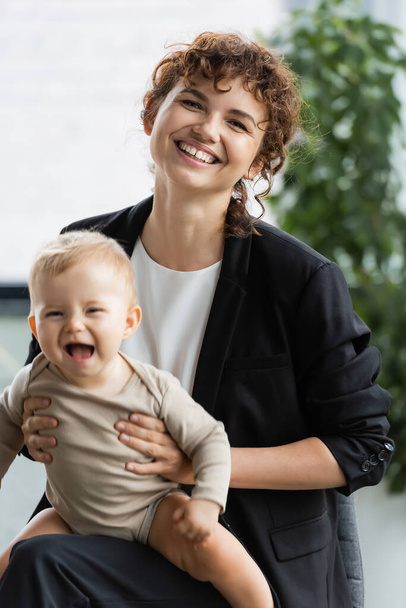 businesswoman in black suit smiling at camera while holding excited baby in office - Photo, image