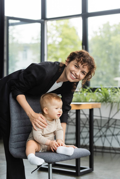cheerful businesswoman smiling at camera near toddler child sitting on office chair - Foto, afbeelding